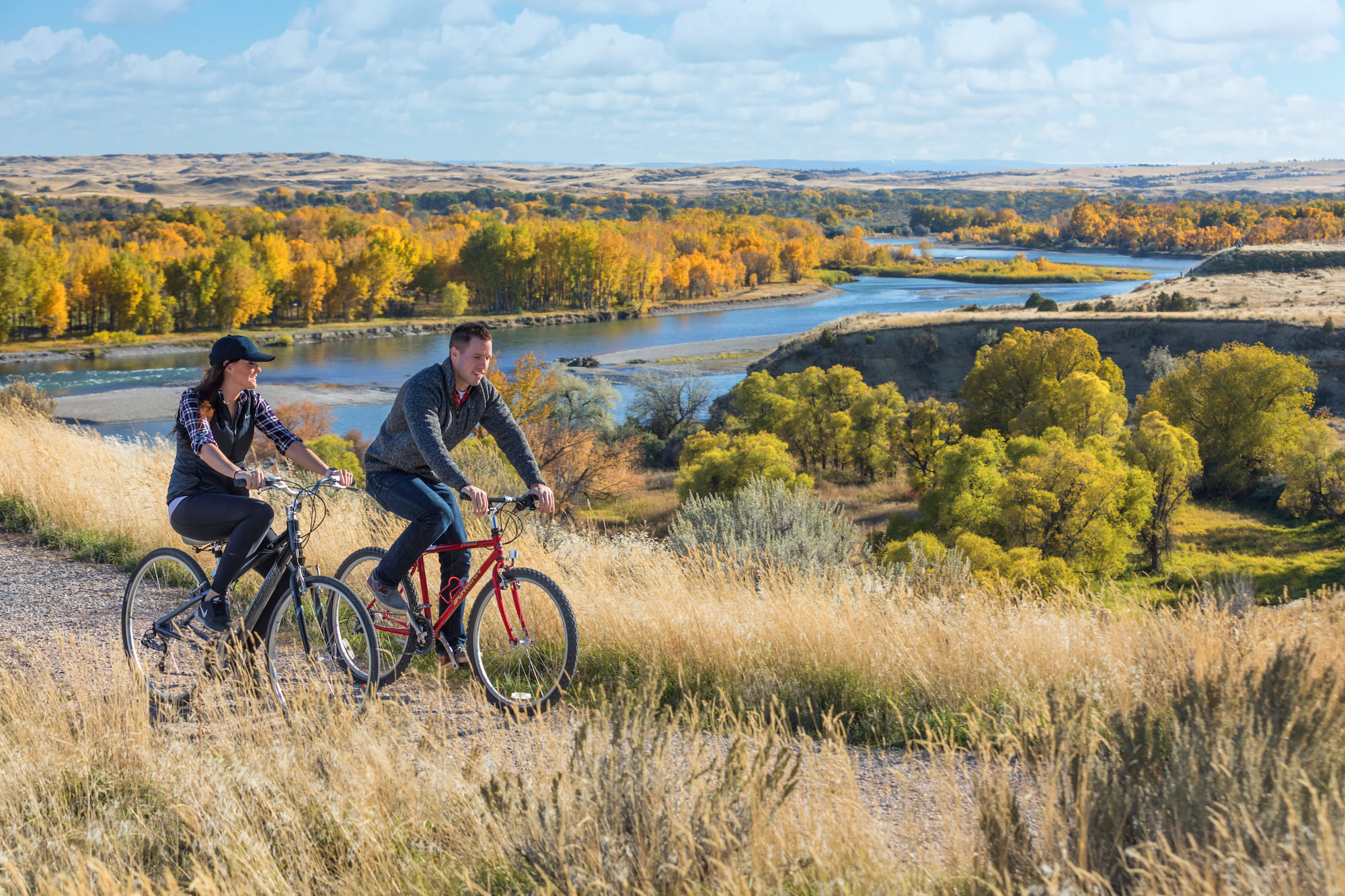 A husband and wife biking along a trail and a river in rural Montana.